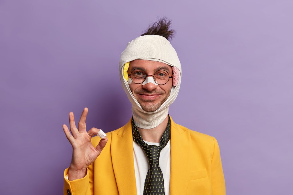 Funny glad man with shiner and broken nose, wears bandage on head, makes okay gesture, assures that everything is okay, smiles pleasantly, forgets about troubles and all his injuries has positive look